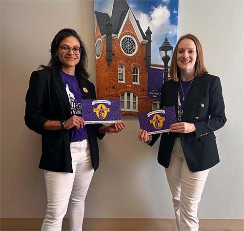 Dean Chandra Hopkins and Jennifer Barksdale ’16 hold up signs to support Rock the Tower in front of a photo of the Wilson Hall tower.